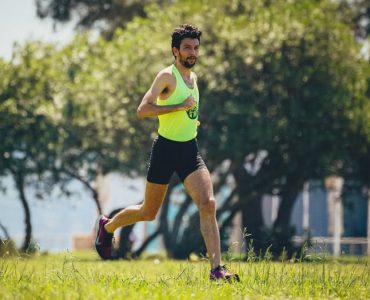 Full length of young bearded sportsman in activewear running on grassy lawn in city park near green trees during outdoor workout