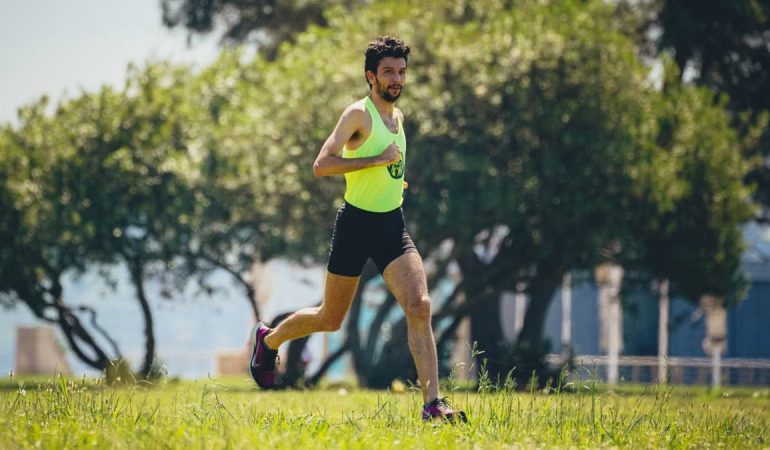 Full length of young bearded sportsman in activewear running on grassy lawn in city park near green trees during outdoor workout